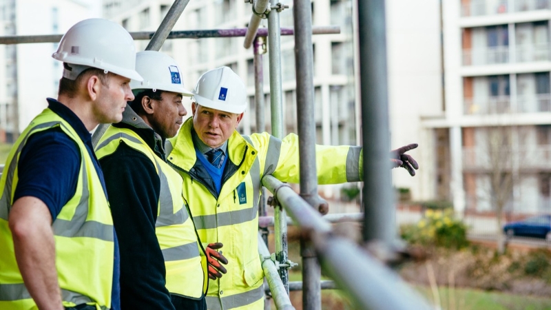 Image shows 3 PPE wearing workers on scaffolding on a construction site. One of the workers is pointing to something off camera