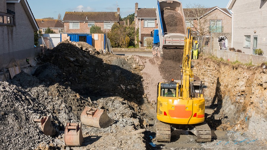 Excavator is digging a house foundation in a residential area while a dumper truck is unloading construction gravel sand and crushed stones on the construction site.